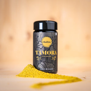 TAMORA Streuwürze BIO in schwarzer Verpackung mit gelbem Aufdruck und Gewürzpulver auf Holzuntergrund.