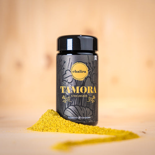 TAMORA Streuwürze BIO in schwarzer Verpackung mit gelbem Aufdruck und Gewürzpulver auf Holzuntergrund.