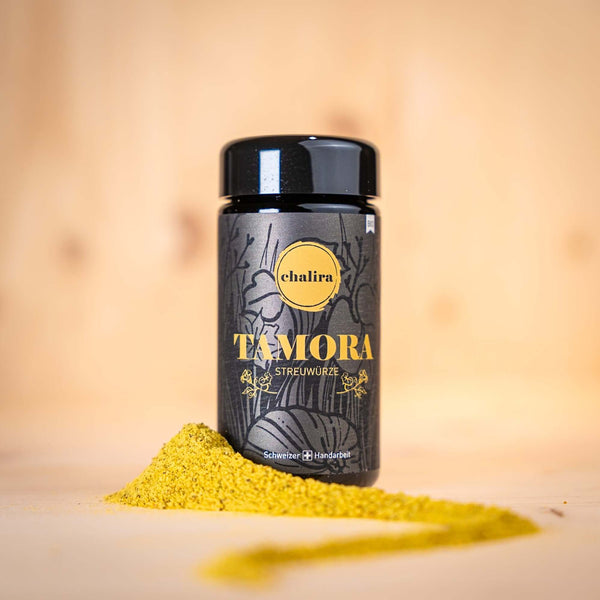 TAMORA Streuwürze BIO in schwarzer Verpackung mit gelbem Aufdruck und Gewürzpulver auf Holzuntergrund.