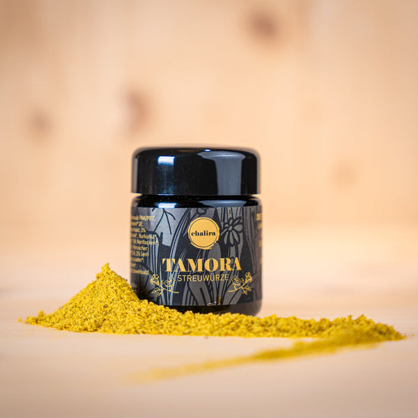 TAMORA BIO Umami-Streuwürze im Glasbehälter mit gelber Gewürzpulver-Haufen auf Holzuntergrund.
